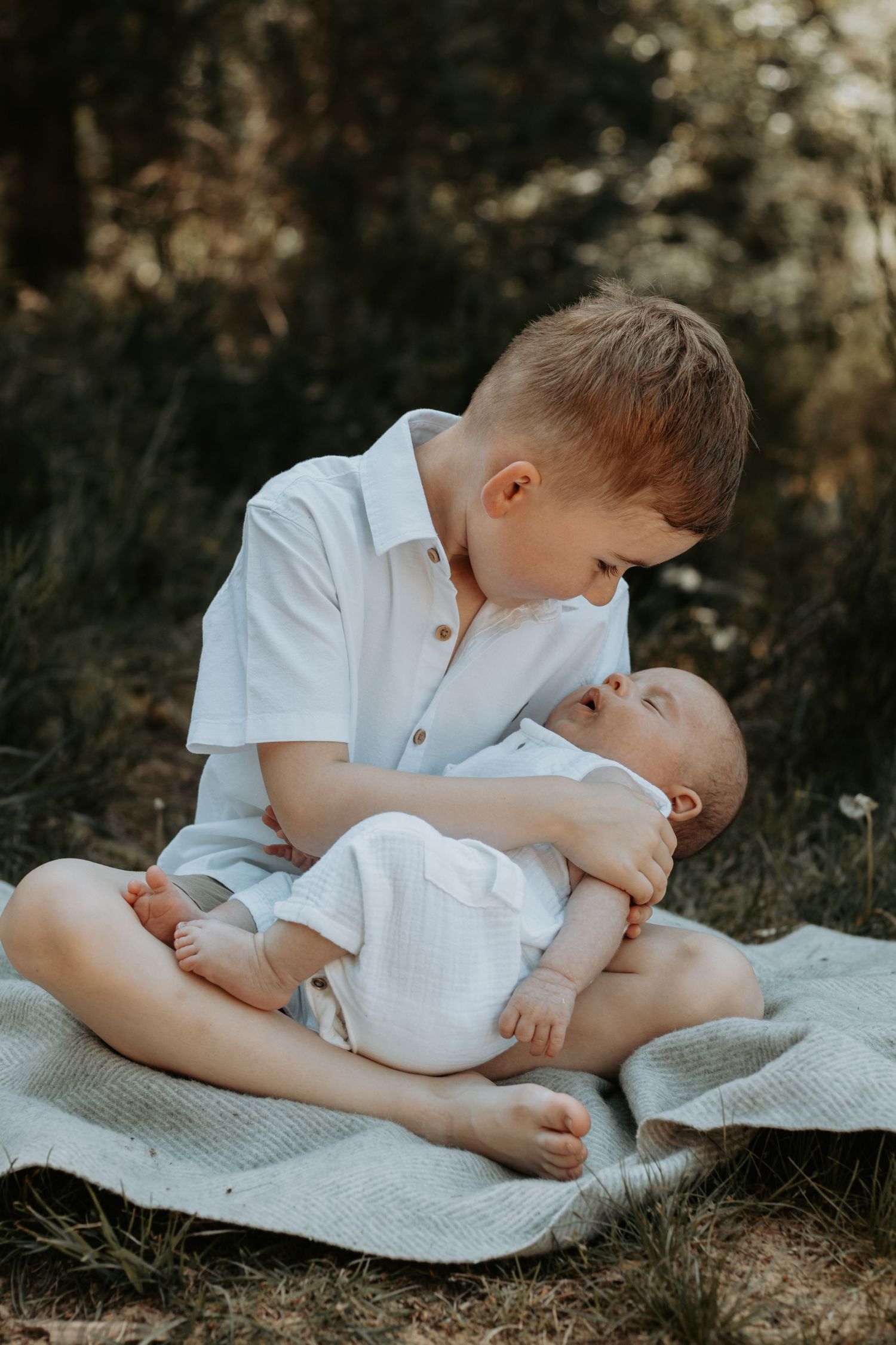 A young boy sits cross-legged outdoors cradling a newborn baby in white clothing on a soft blanket.
