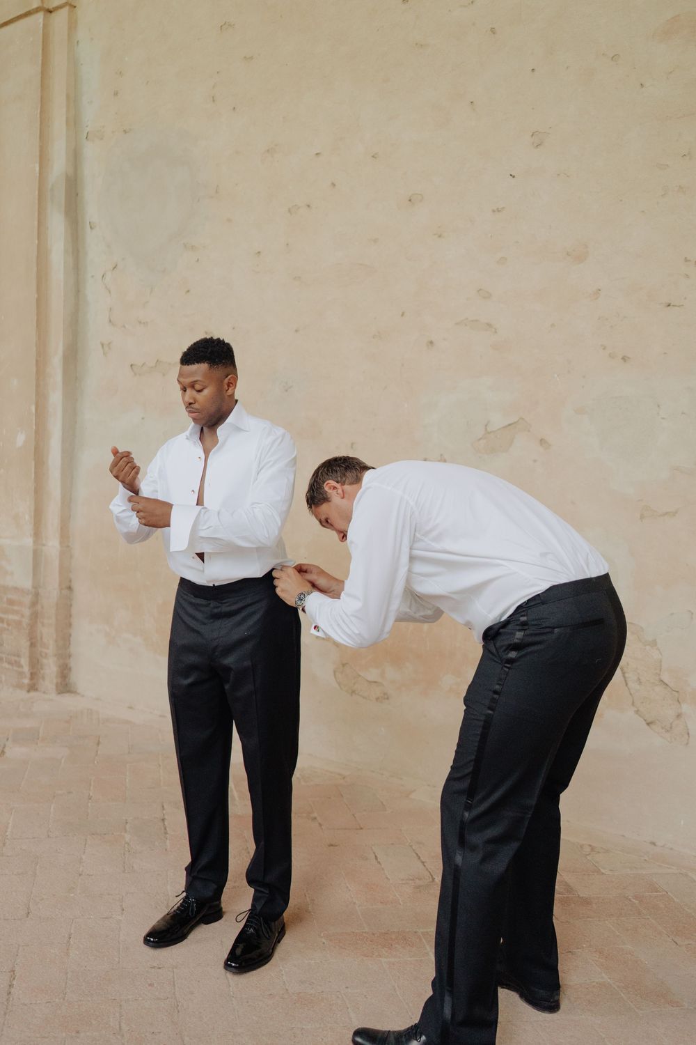 Groomsmen in formal attire perform synchronized dance moves against a beige wall.