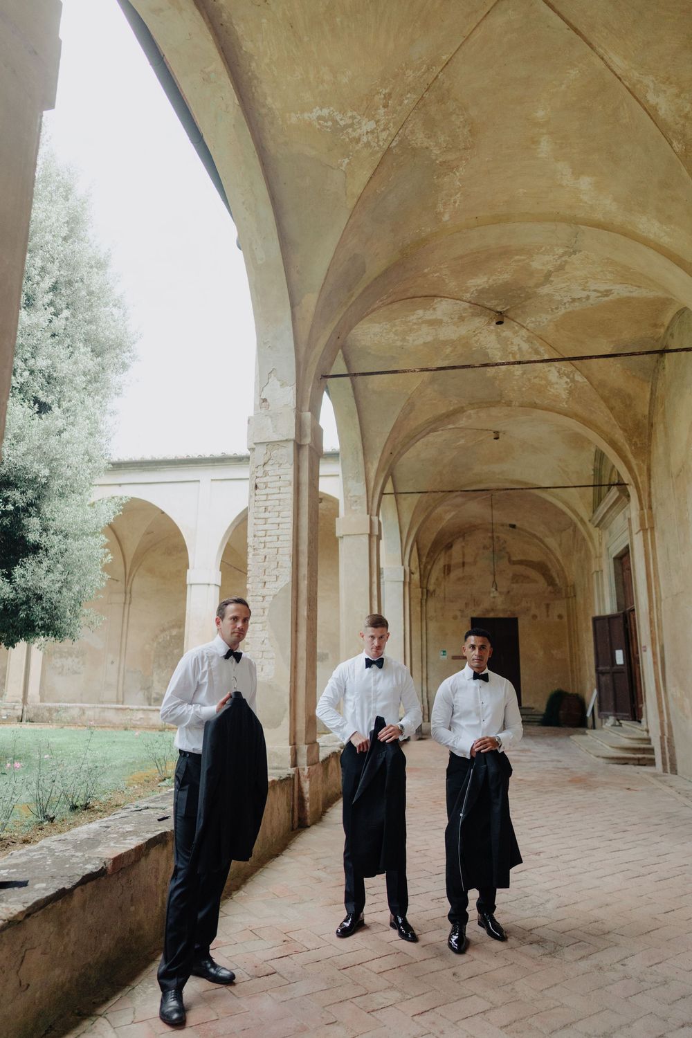 Three waiters in formal attire stand beneath historic arched corridors of an Italian villa.