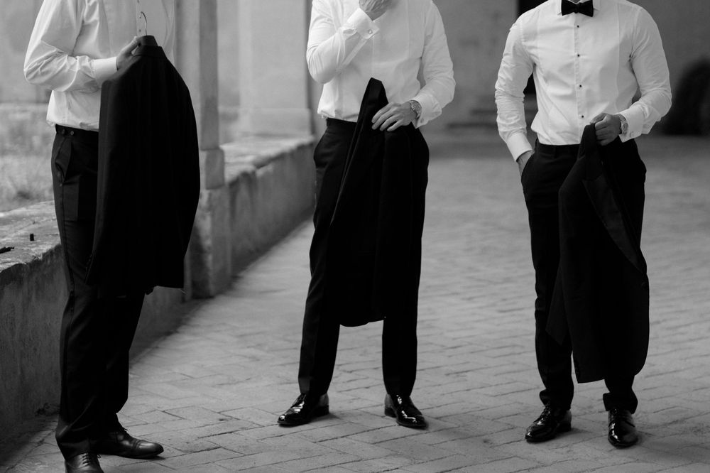 Three men in formal attire holding suit jackets in a monochrome hallway.