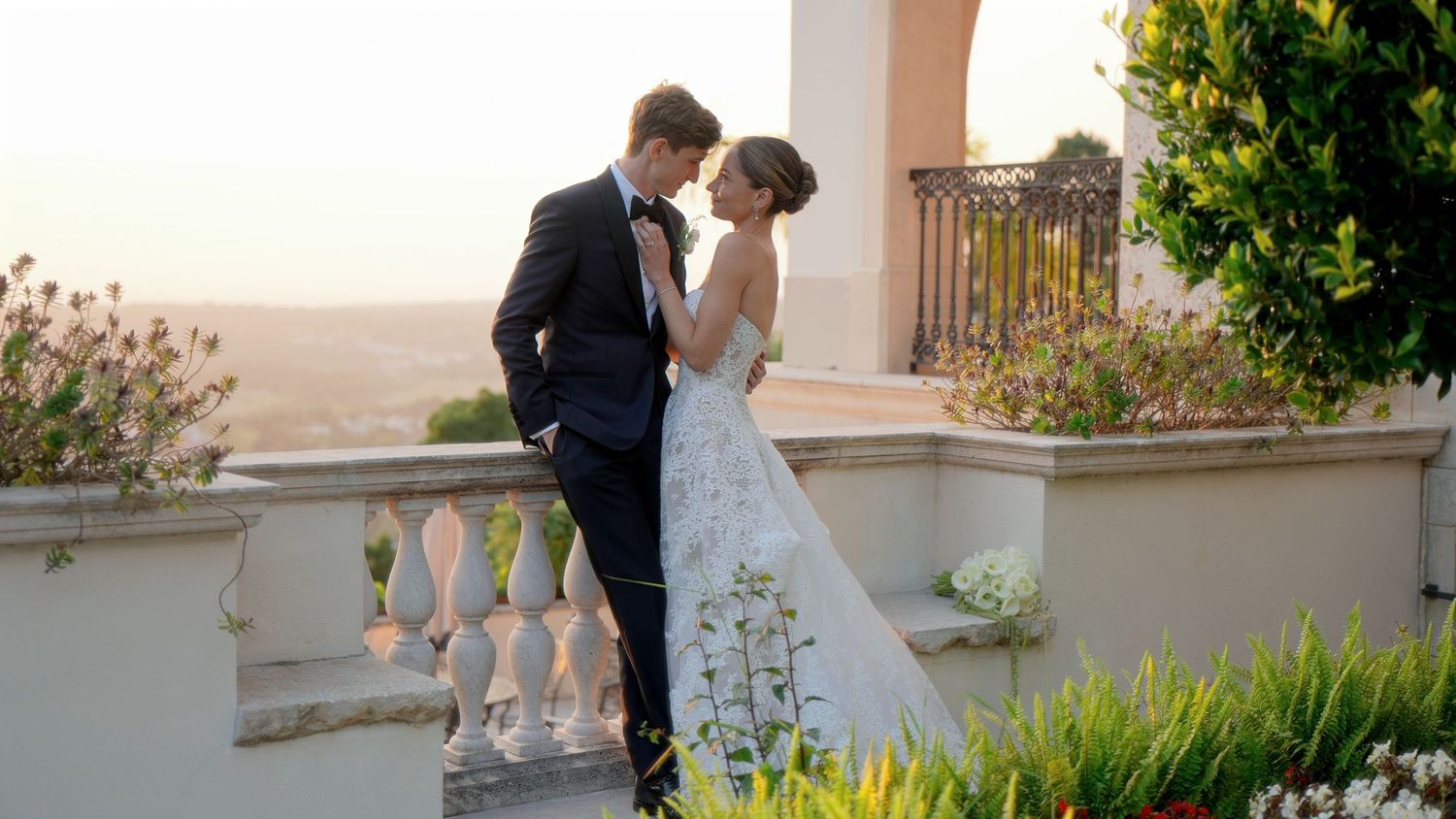 A couple walks together along a beige stucco wall with lush landscaping and flowering plants in an outdoor setting.