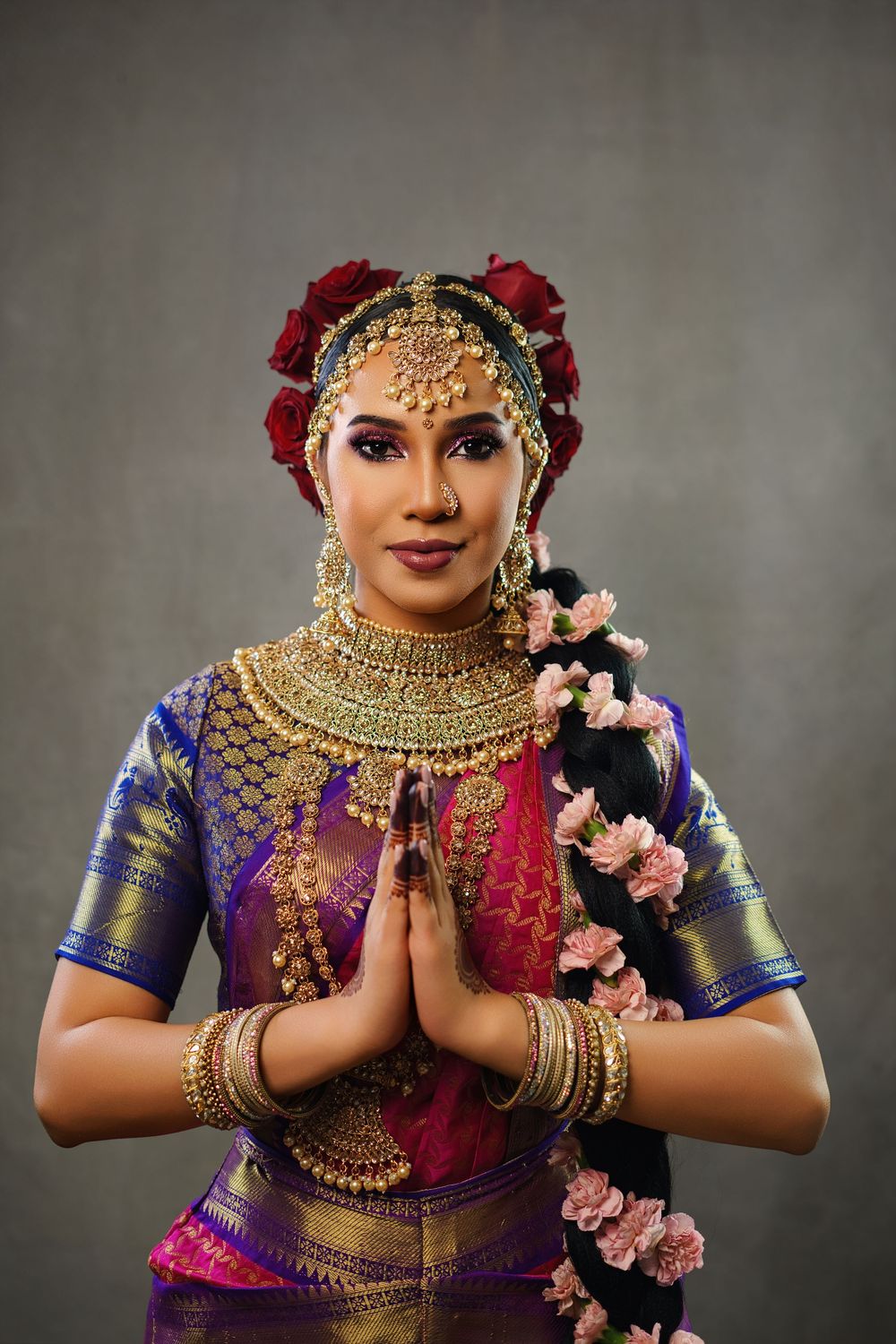 A traditional Indian dancer in vibrant purple and pink attire with gold jewelry performs a graceful namaste gesture.