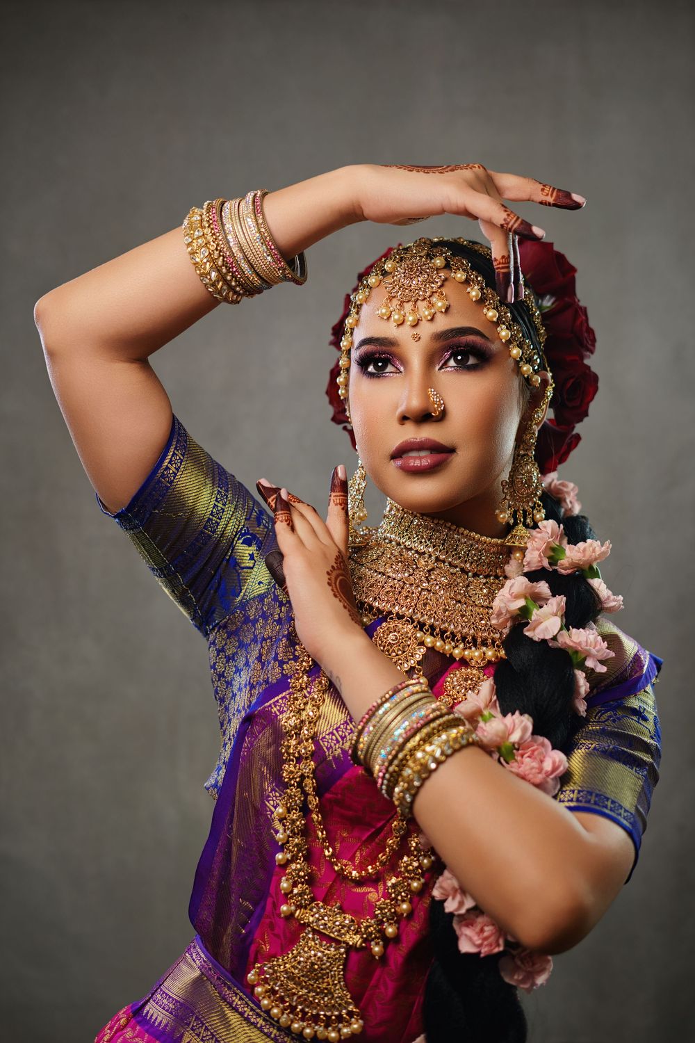 Traditional Indian dancer in ornate gold jewelry and purple silk sari strikes graceful pose with red roses in hair.