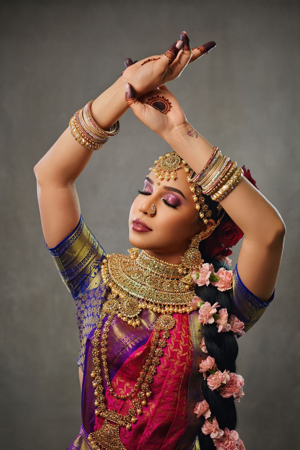 Traditional Indian dancer in purple and pink attire with ornate gold jewelry performs graceful hand movements.