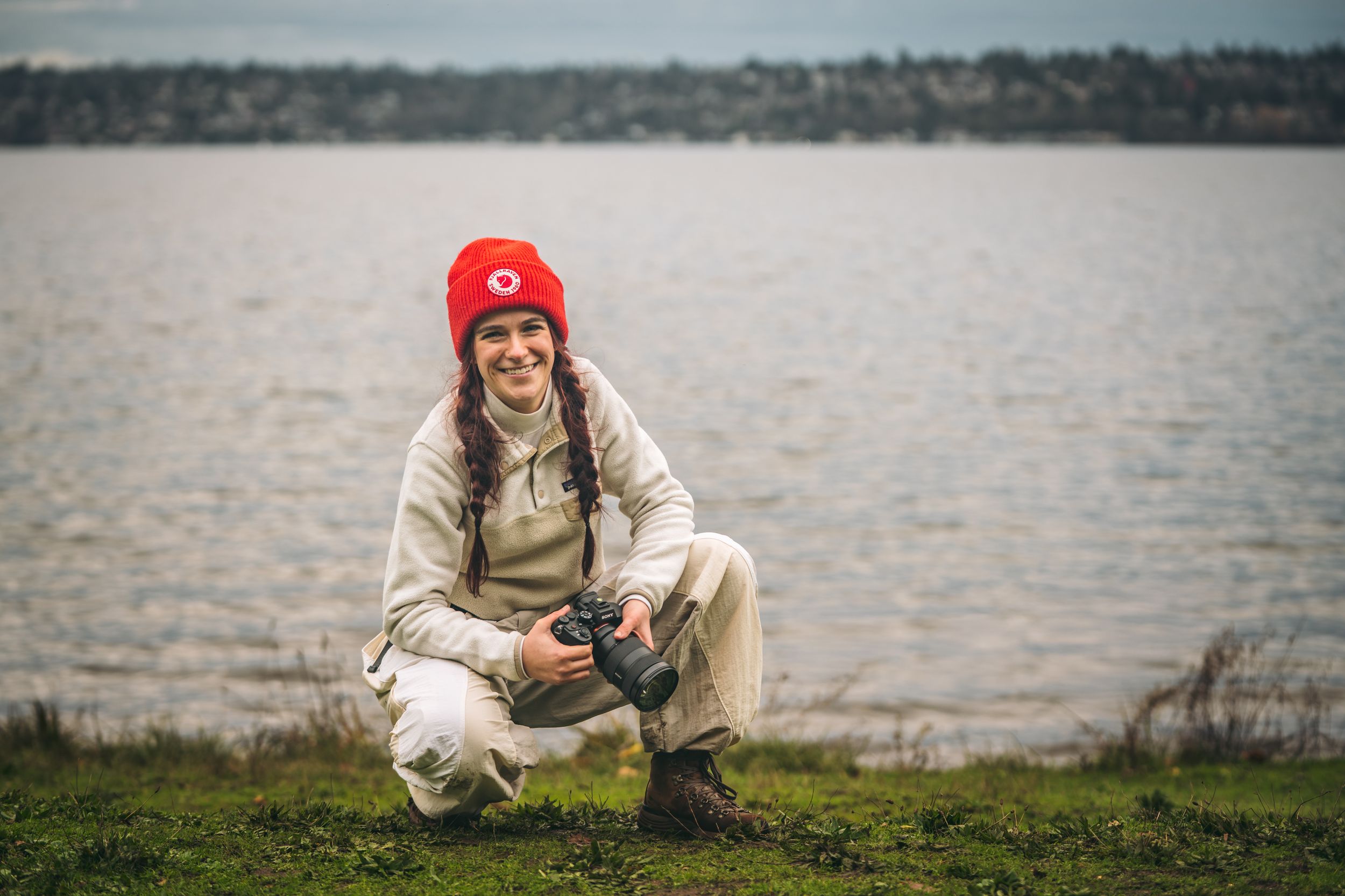 Smiling woman in red beanie crouching with a camera near a calm lake shore.