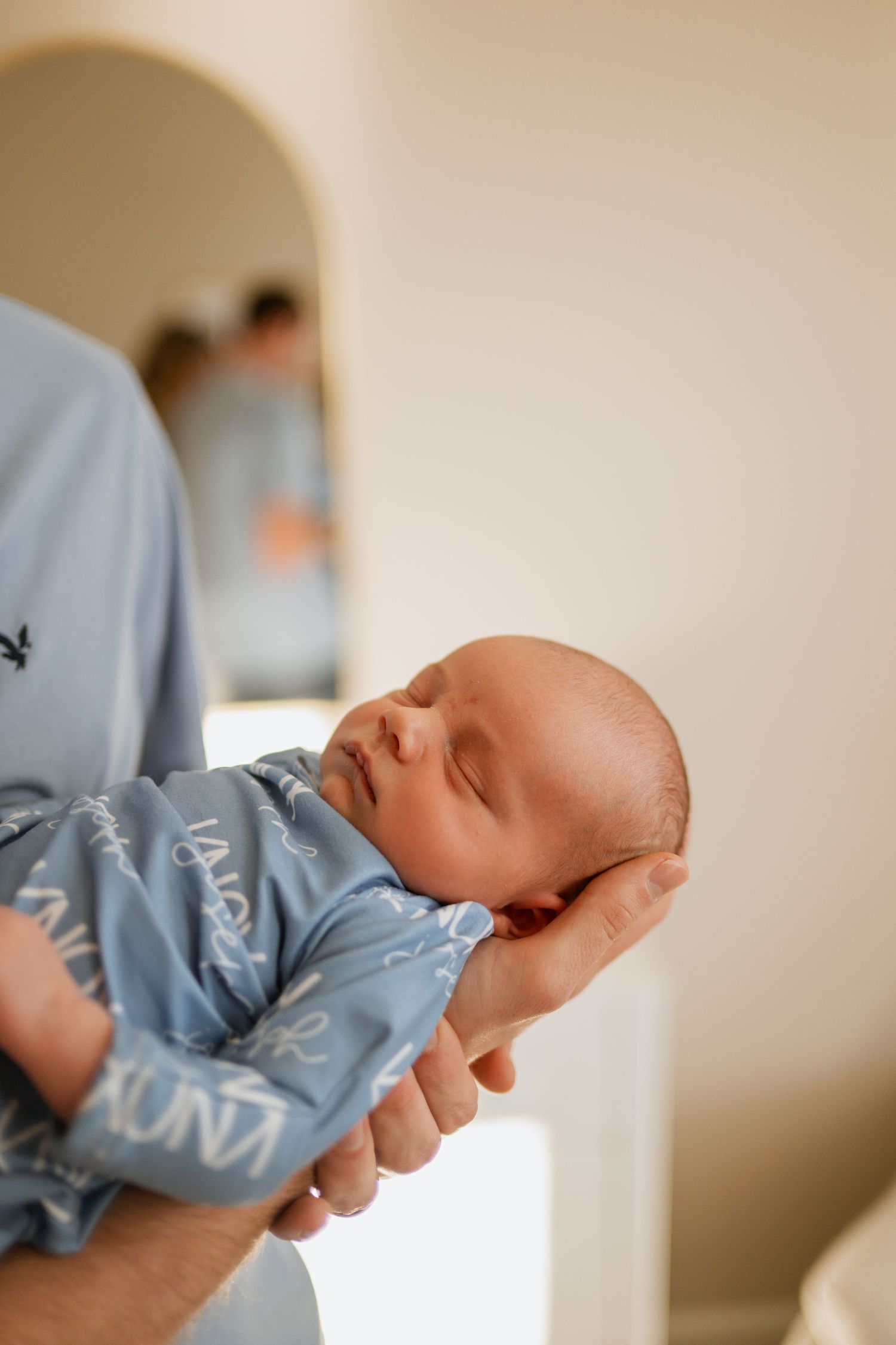 A sleeping newborn baby wearing a light blue onesie rests peacefully in a caregiver's arms.