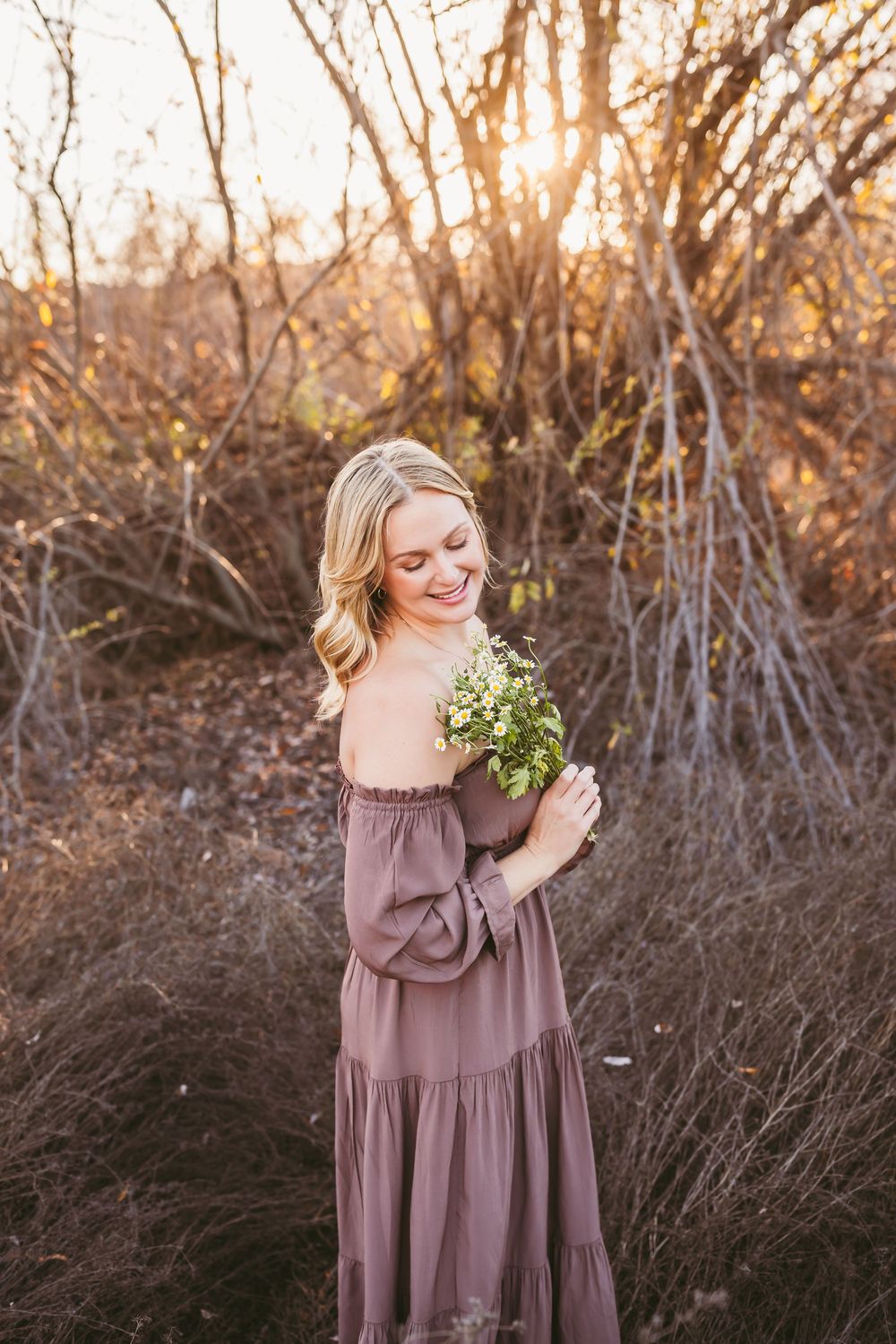 A person in a flowing mauve dress holds wildflowers while standing in tall grass during golden hour.