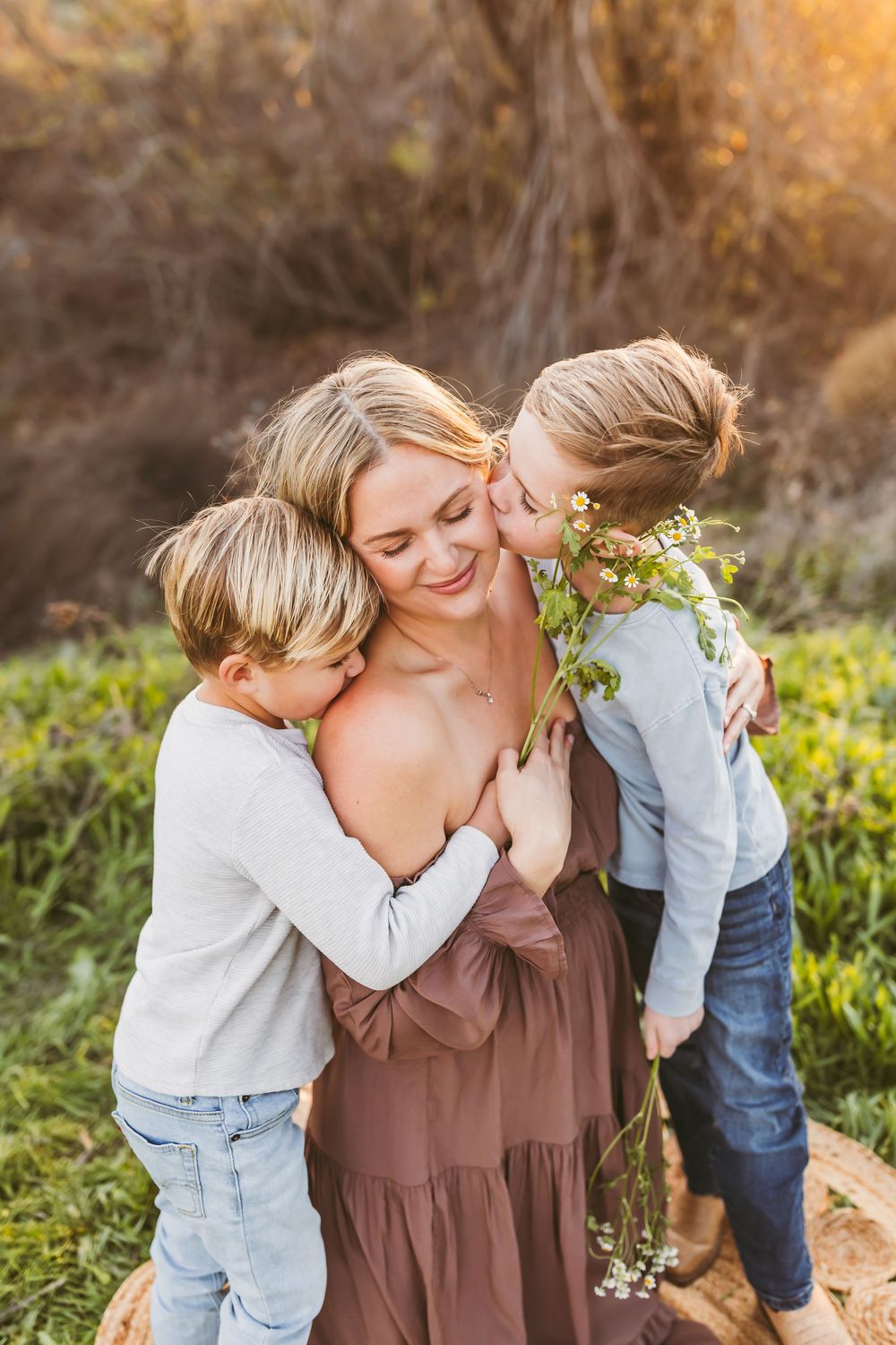 A tender outdoor family moment captured in warm golden light among wildflowers and natural scenery.