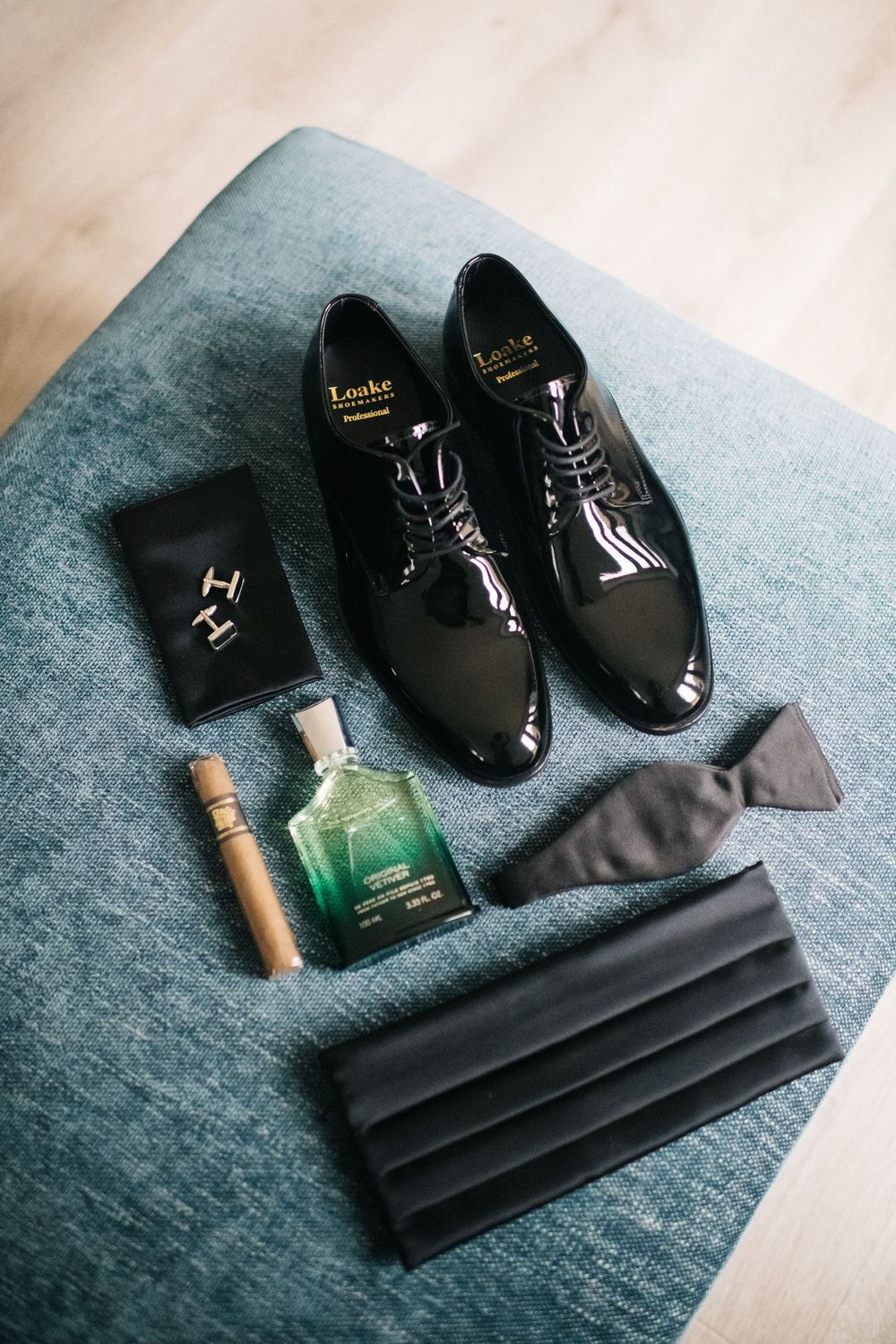 Groom's wedding accessories including black dress shoes, tie, and cufflinks on gray fabric.