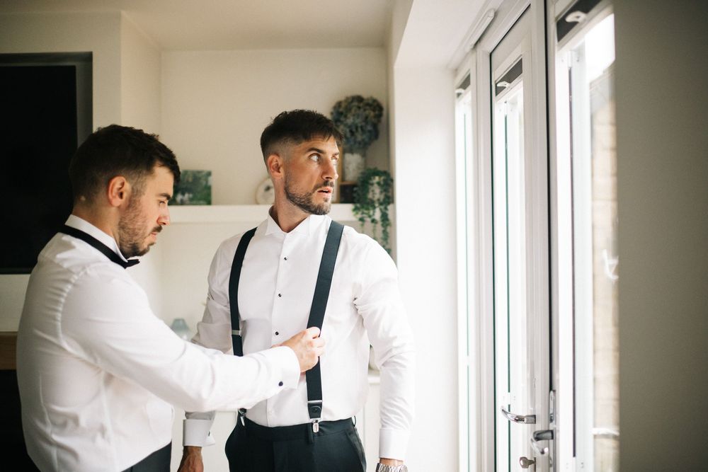 Two groomsmen in white shirts and suspenders helping each other get ready.