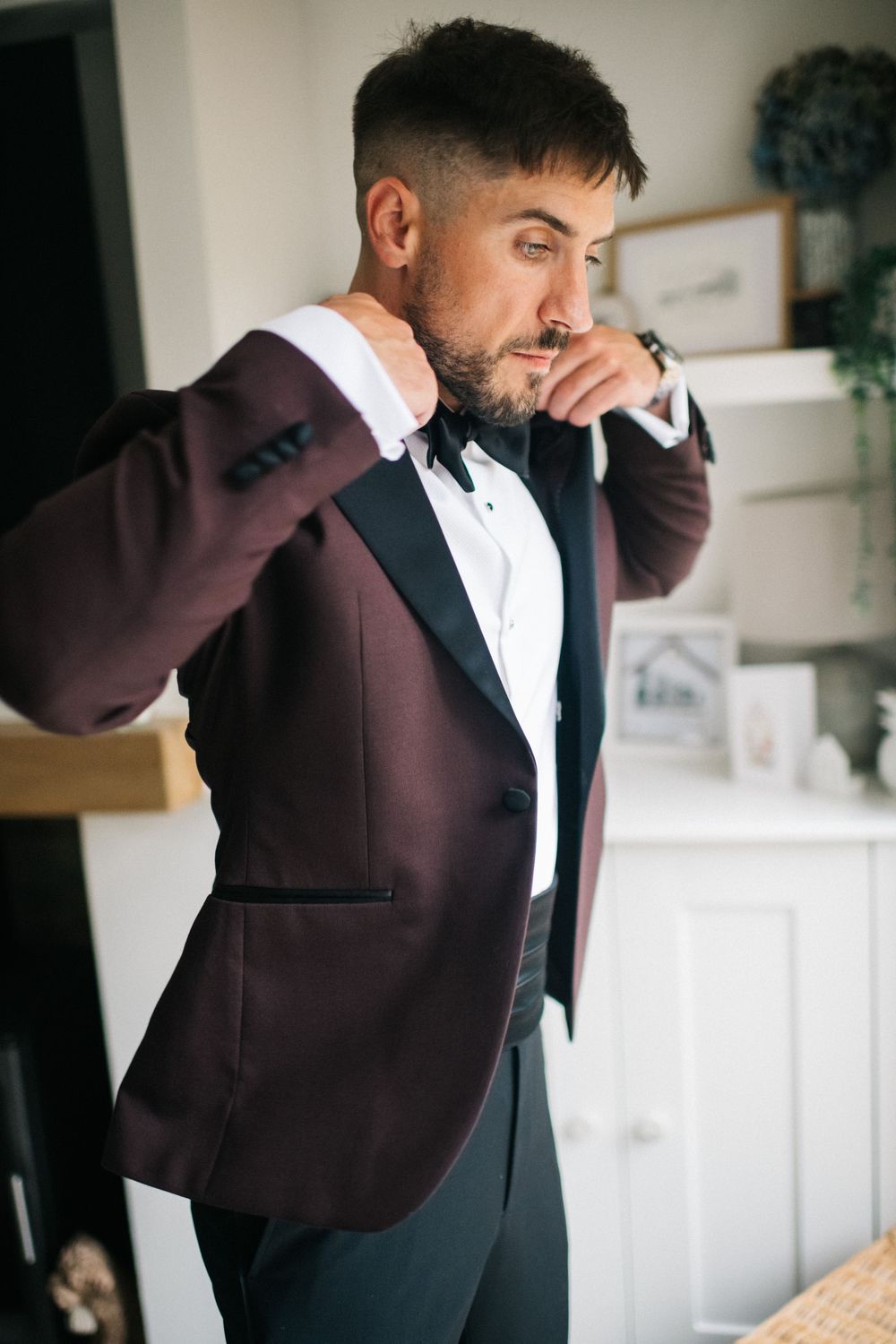 Groom adjusts his burgundy tuxedo jacket while getting ready for wedding.