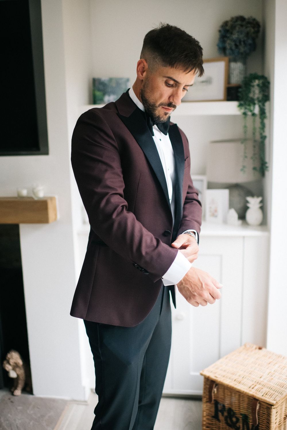 Groom in burgundy suit jacket adjusts cufflinks during wedding preparation.