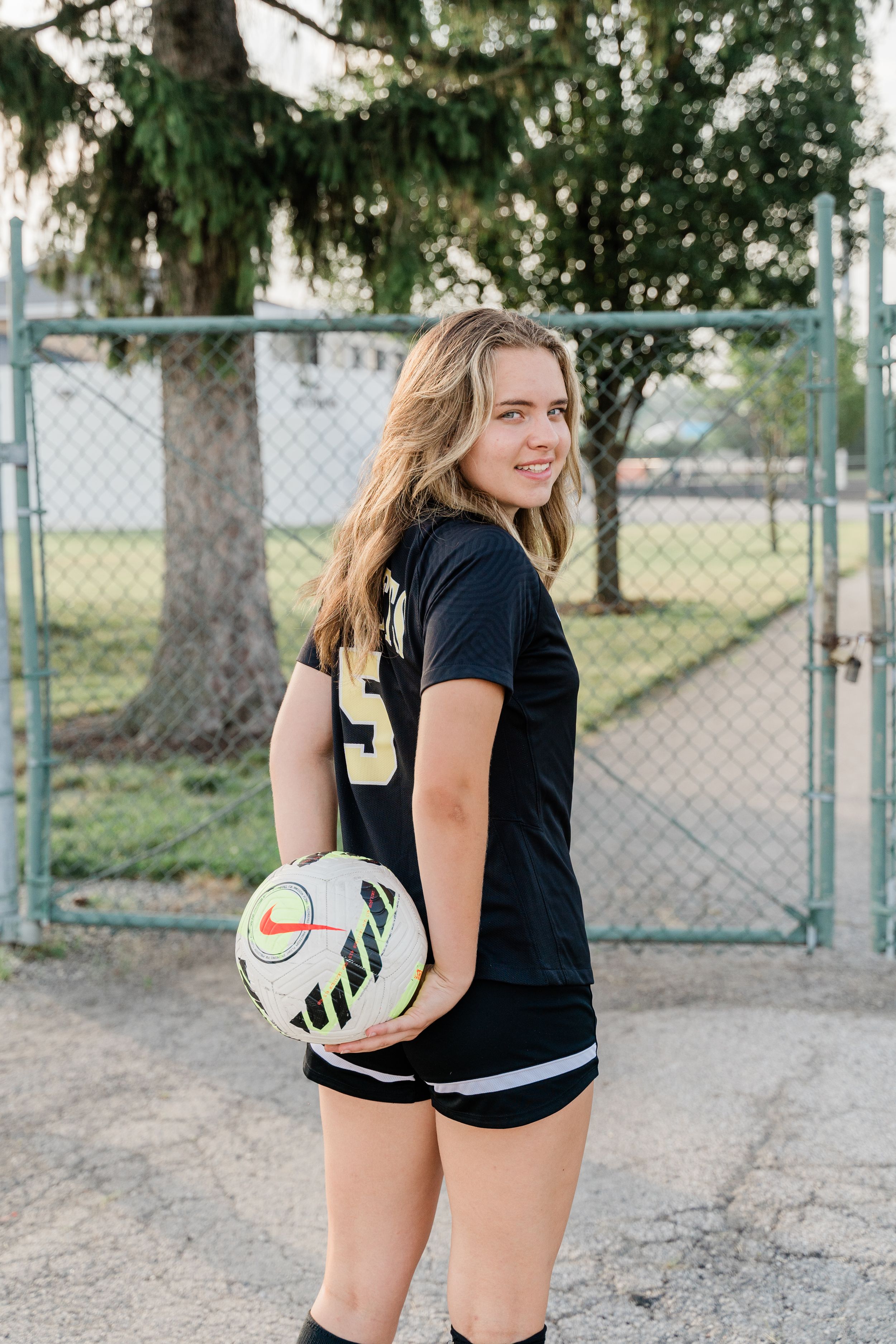 Athlete in black sportswear holding a volleyball on outdoor court with trees in background.