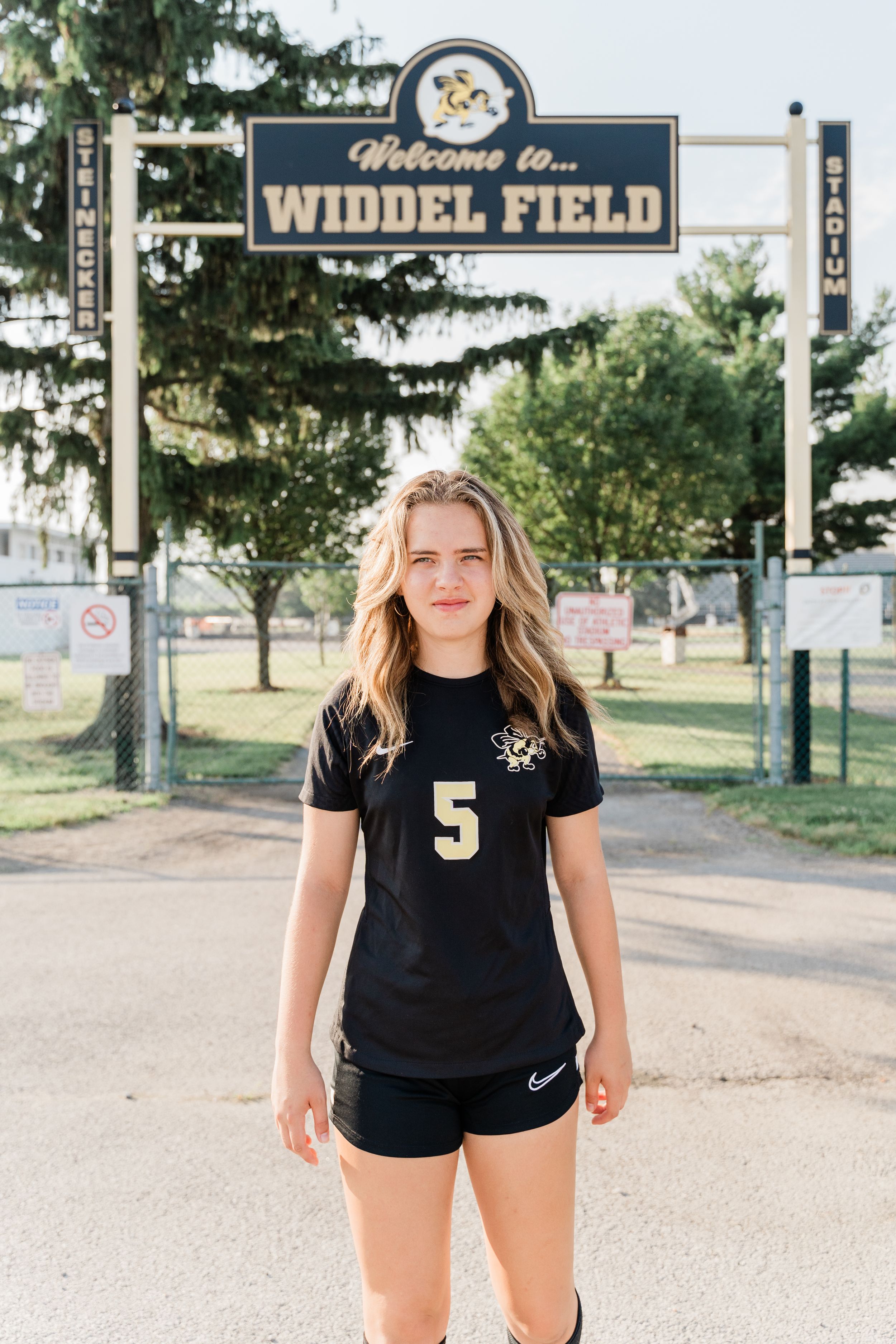 Soccer player stands in front of Widdel Field sign wearing black jersey number 5 and shorts on sunny day.