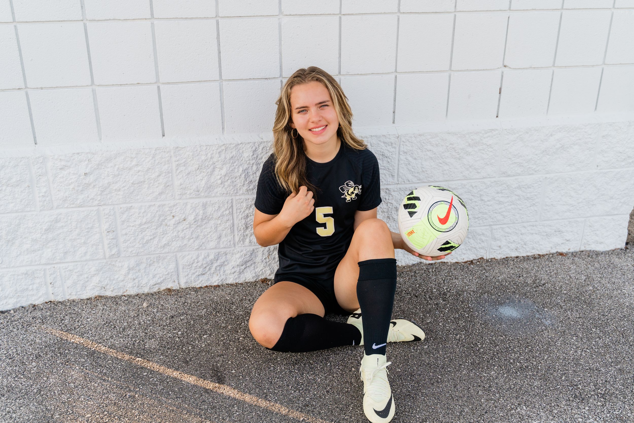 A person wearing a black t-shirt gives a thumbs up while sitting on the ground next to a white soccer ball.