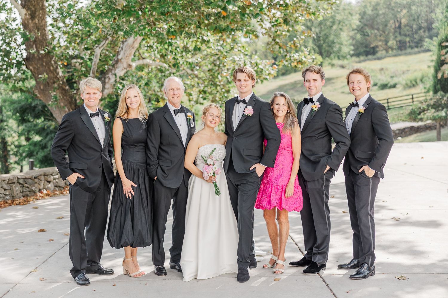 Wedding party in formal attire poses together outdoors with black dresses and tuxedos.