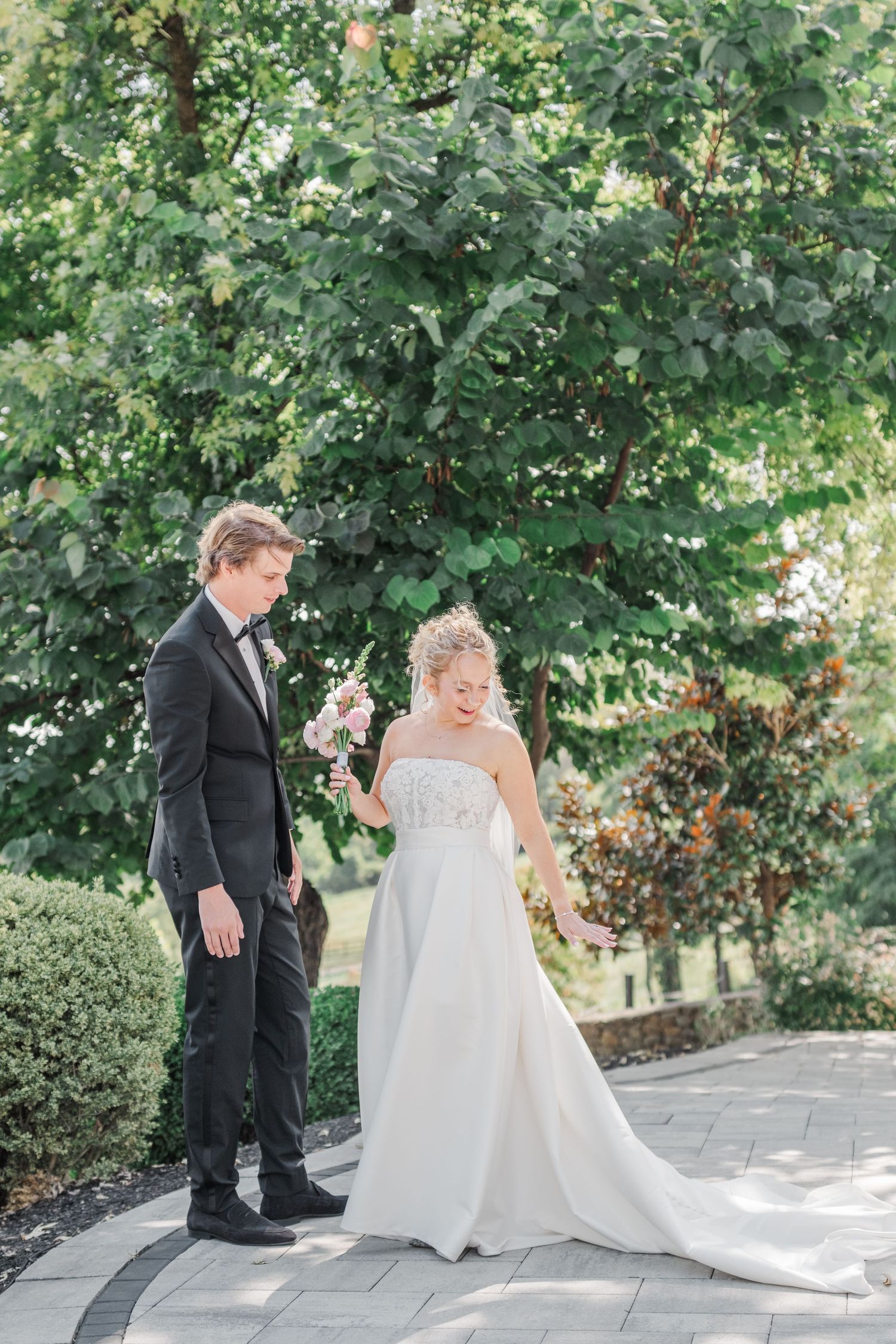A couple in wedding attire stands on a stone path surrounded by lush green trees and landscaping.