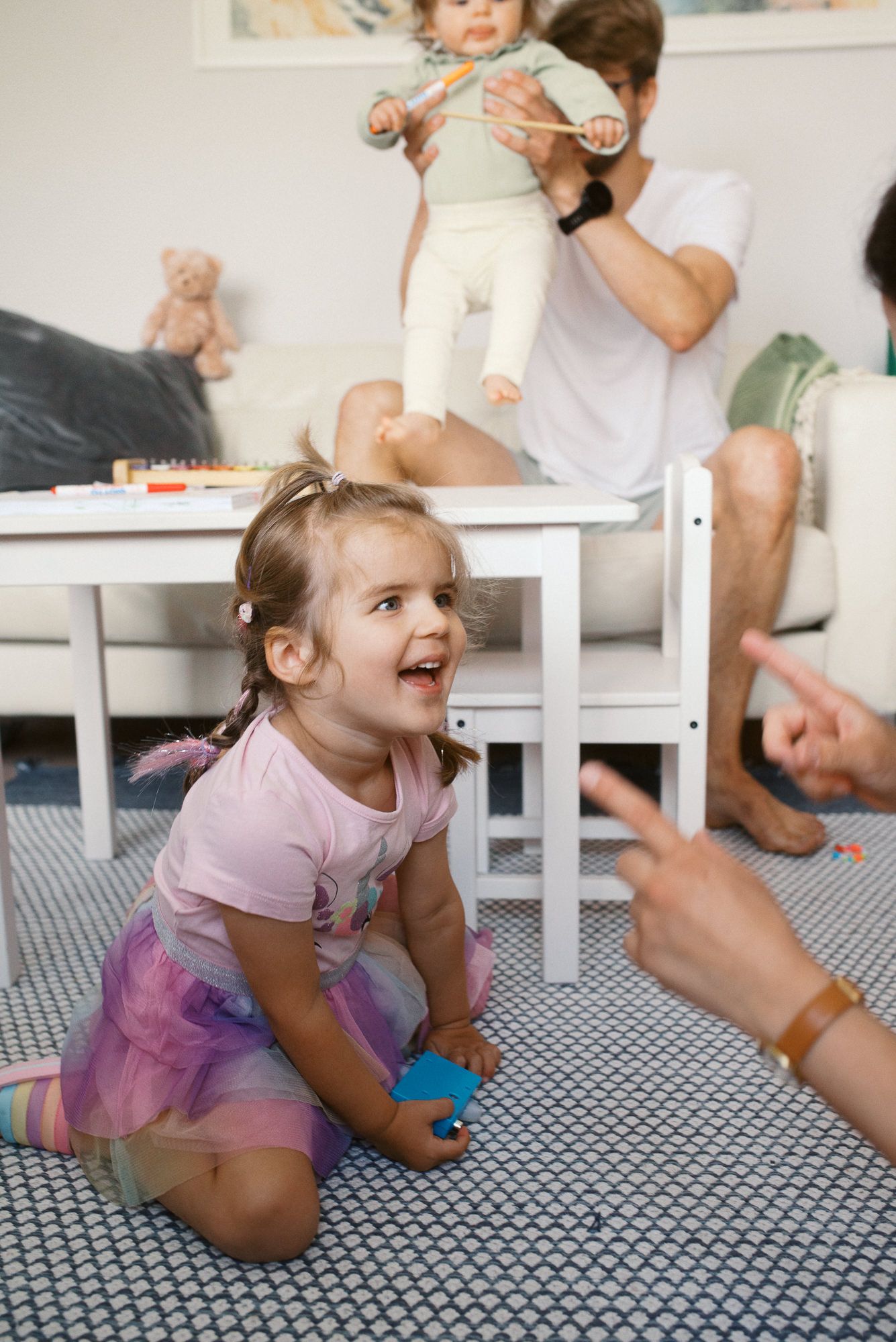 Young child in pink shirt and tulle skirt plays on floor of living room while family gathers around.