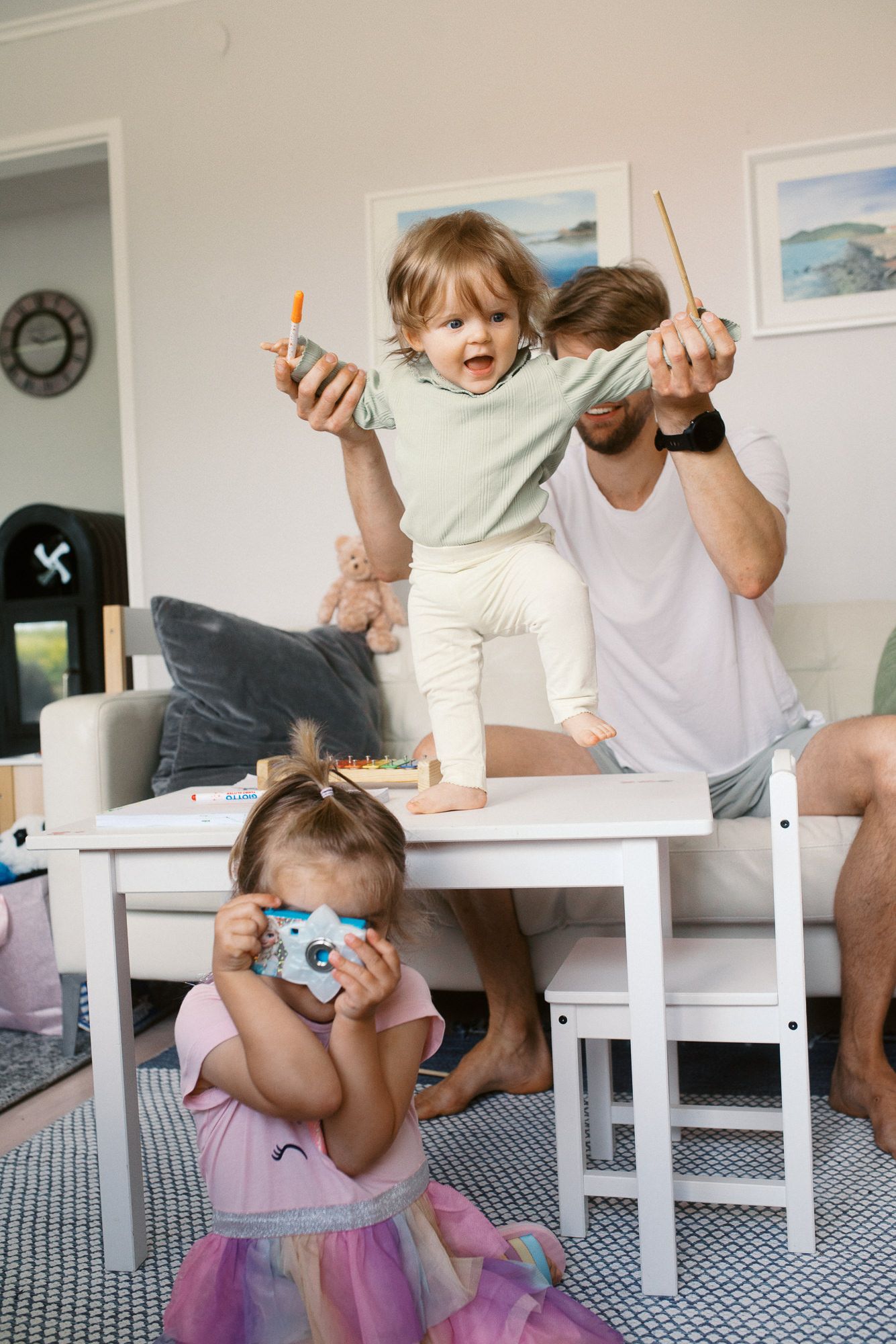 A playful family moment at home with children on a small white table while someone takes photos with a camera.