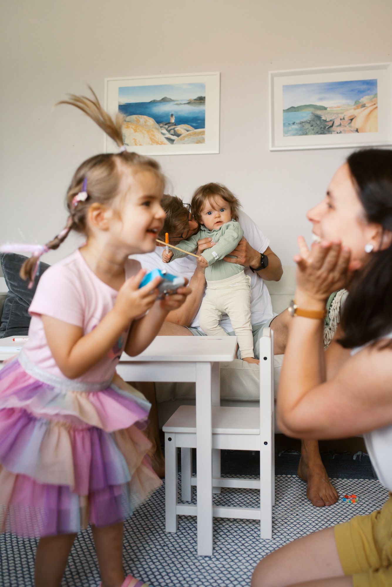 A playful scene in a room with coastal artwork, featuring a tulle dress, toys and joyful interaction on a patterned rug.