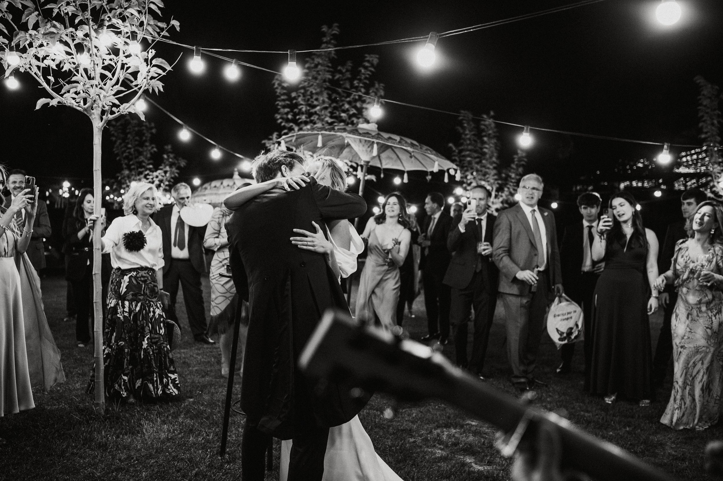 Wedding guests dance and celebrate under string lights at an outdoor evening reception.