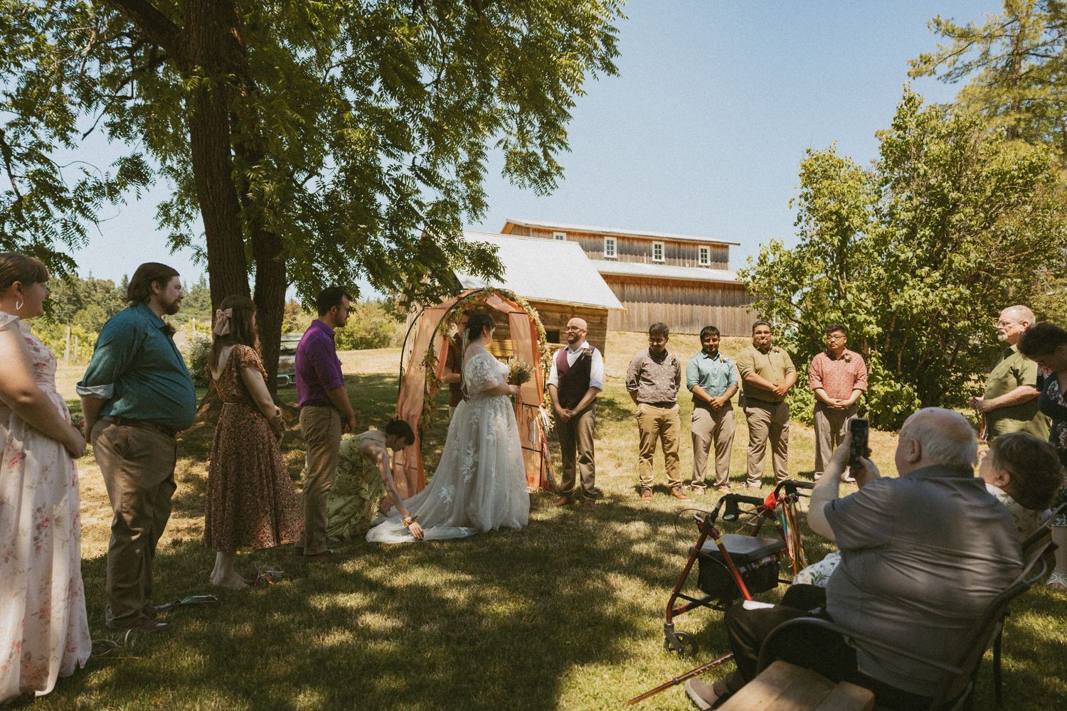 A group of wedding guests gather outdoors under trees during a sunny afternoon celebration.