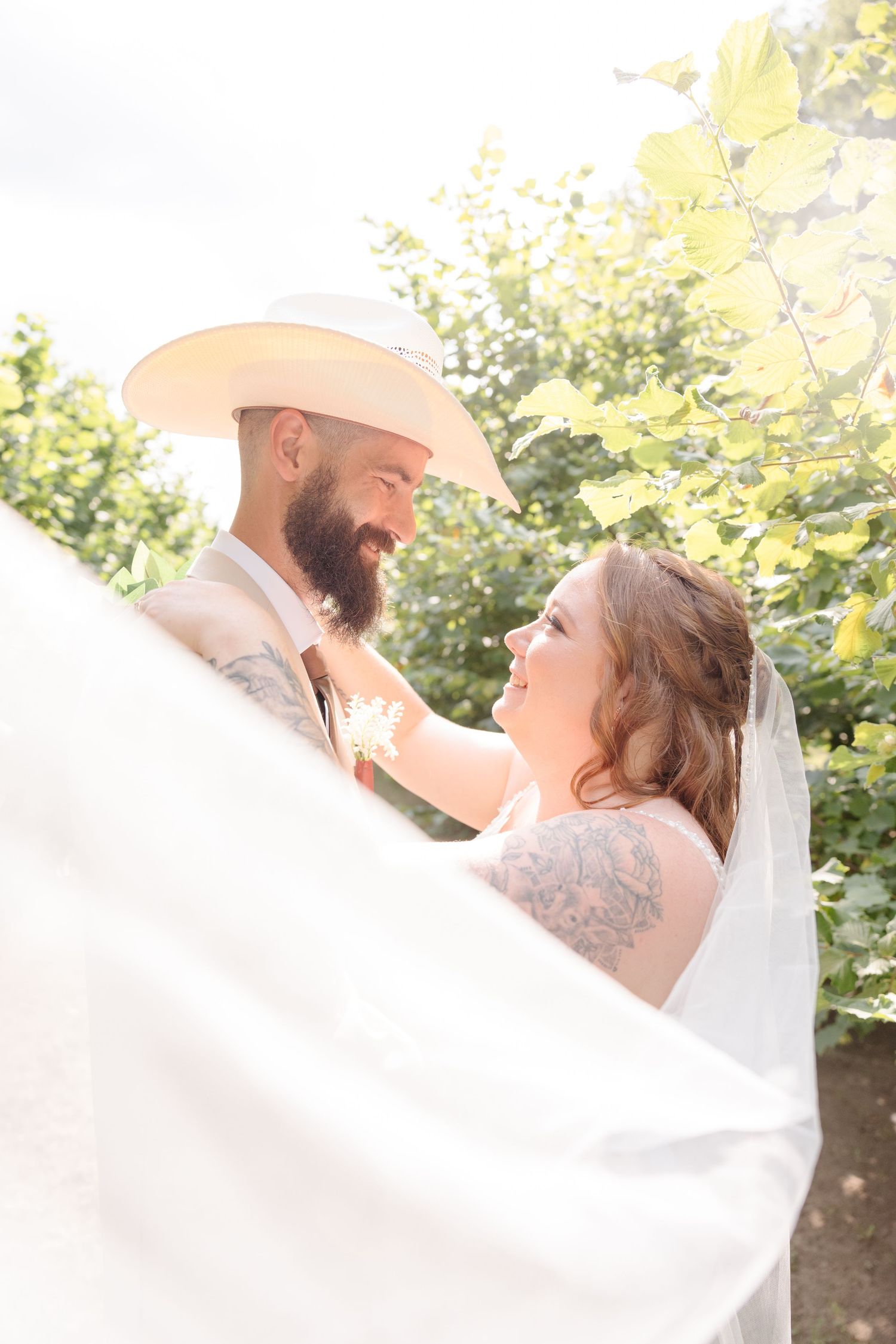 A romantic wedding couple shares an intimate moment outdoors with a flowing white veil in the sunlight.