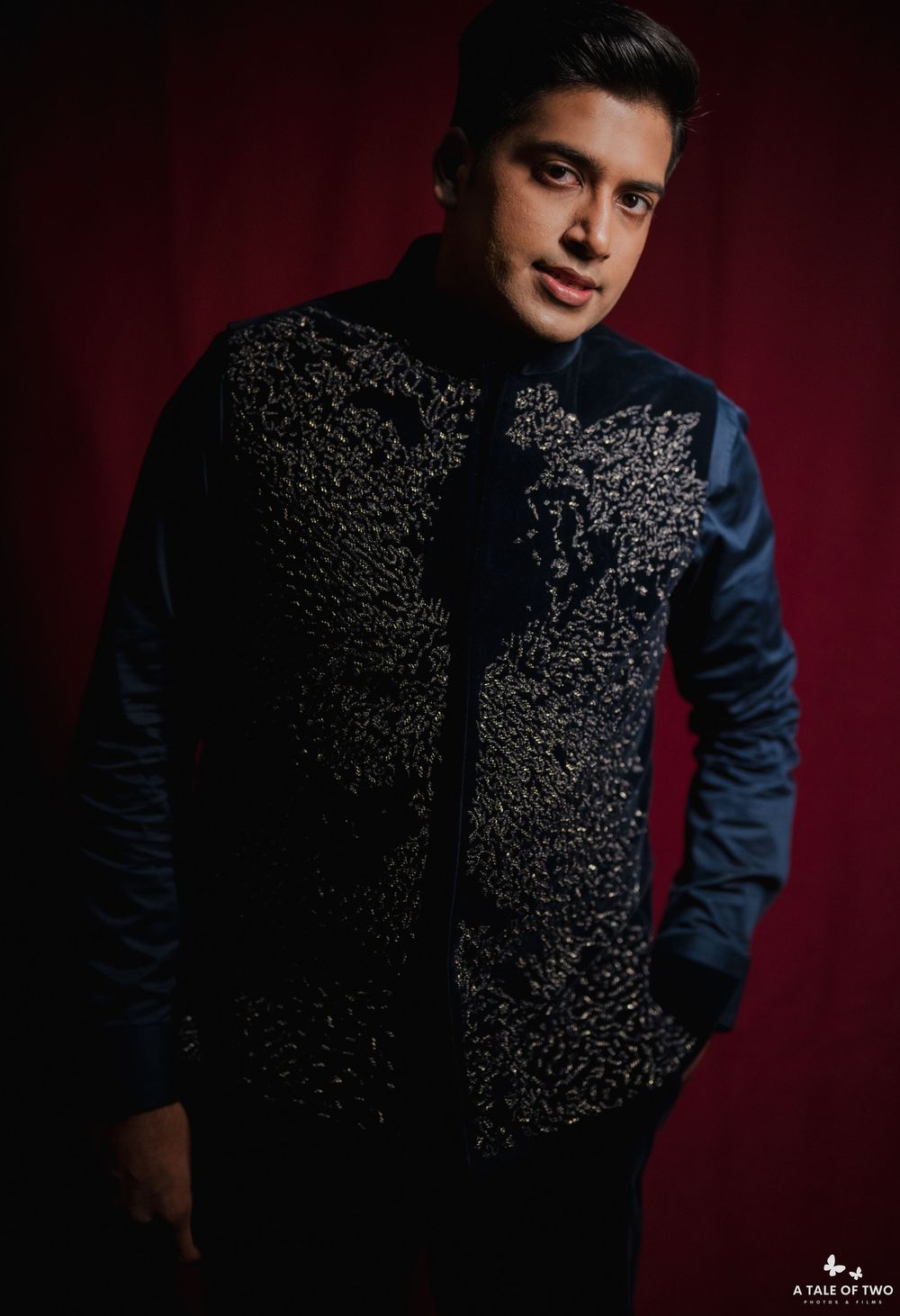 Professional studio portrait against dark red backdrop featuring person wearing navy suit with intricate black textured pattern.