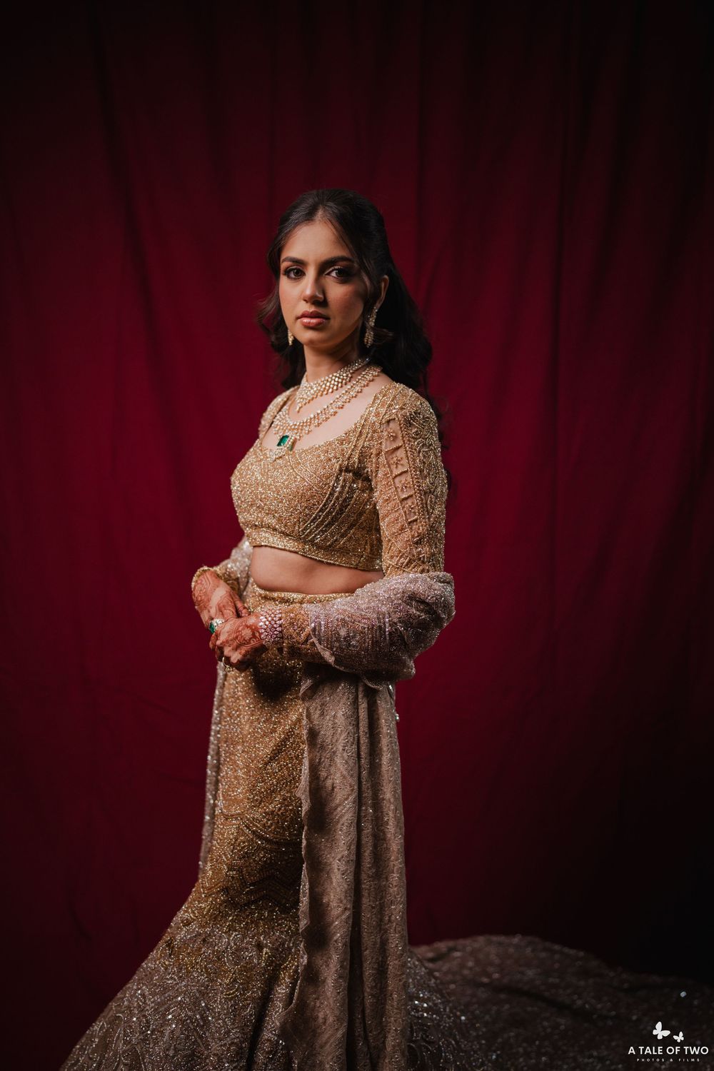 A figure in a gold embellished traditional Indian lehenga poses against a deep red velvet backdrop in a maternity photoshoot.