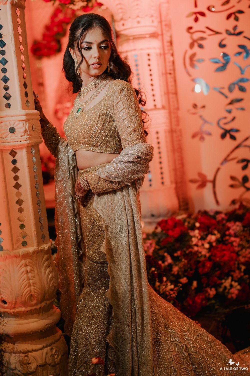 Elegant figure poses in a beige embroidered gown against ornate coral-colored pillars and floral decorations.