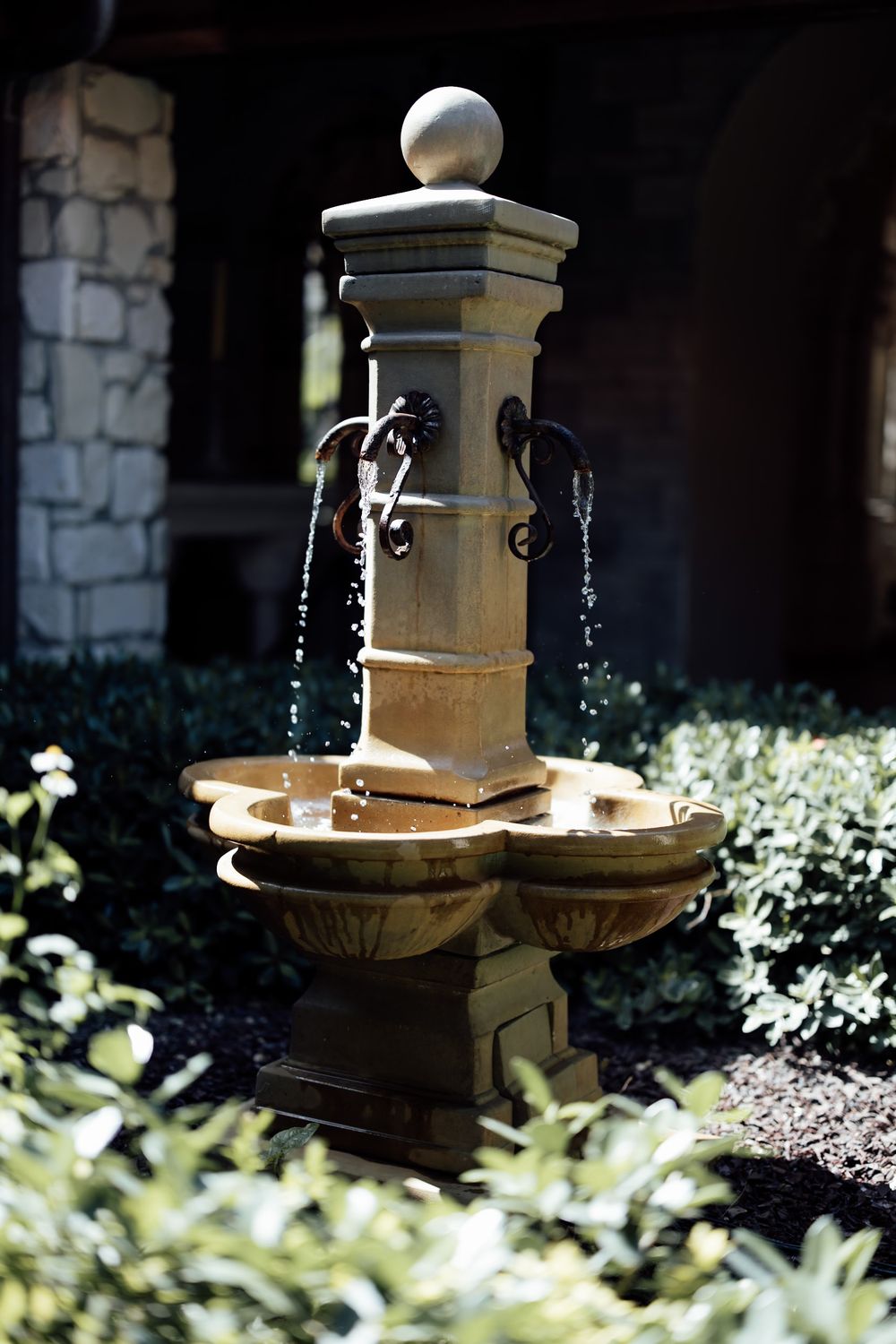 Stone fountain with decorative tiers in a garden setting with sunlit foliage.