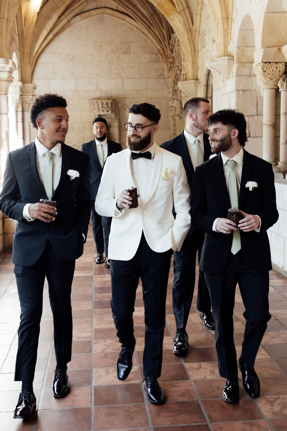 Groomsmen in black tuxedos and white ties walking in formation through an elegant archway.