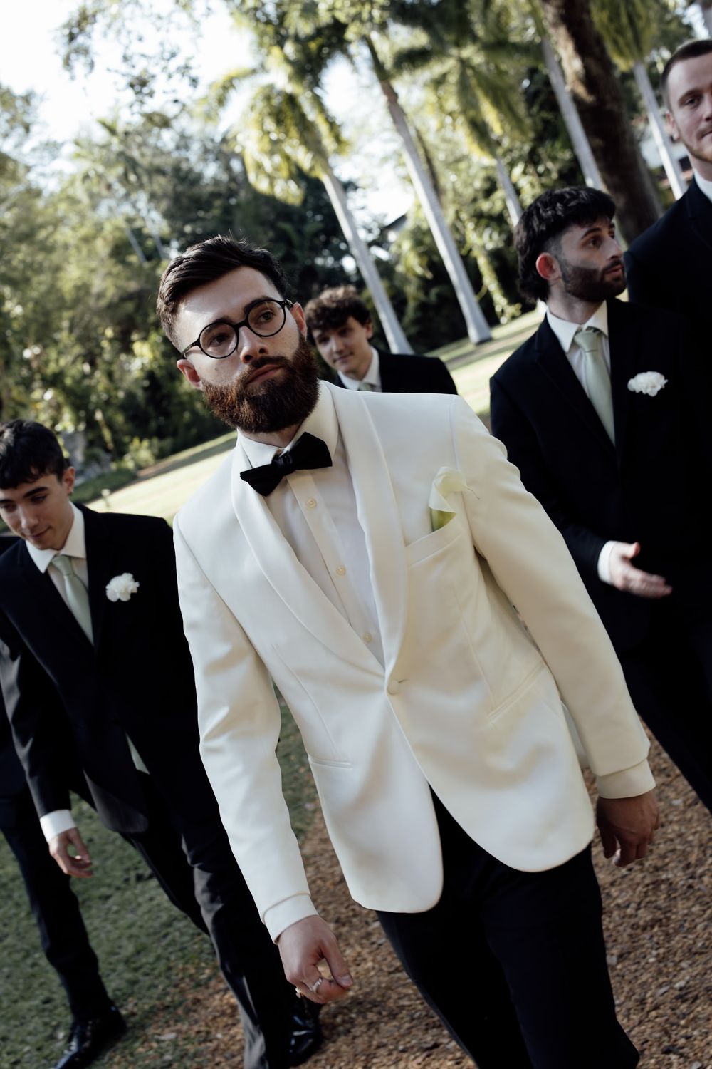Groomsmen in black suits and white tuxedo jackets gather for wedding photos under palm trees.