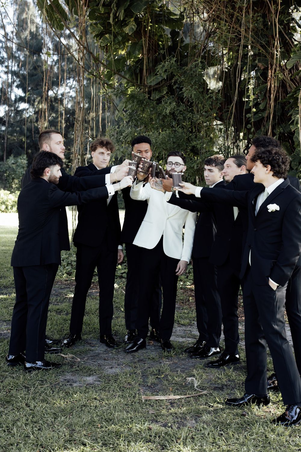 Wedding party in formal attire gathered outdoors among tall trees for group photos.
