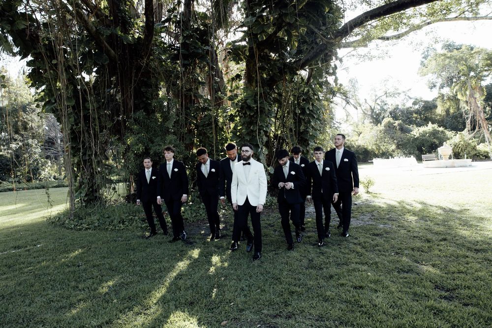 Groomsmen in black suits and one in white walk together on a grassy lawn during an outdoor wedding.
