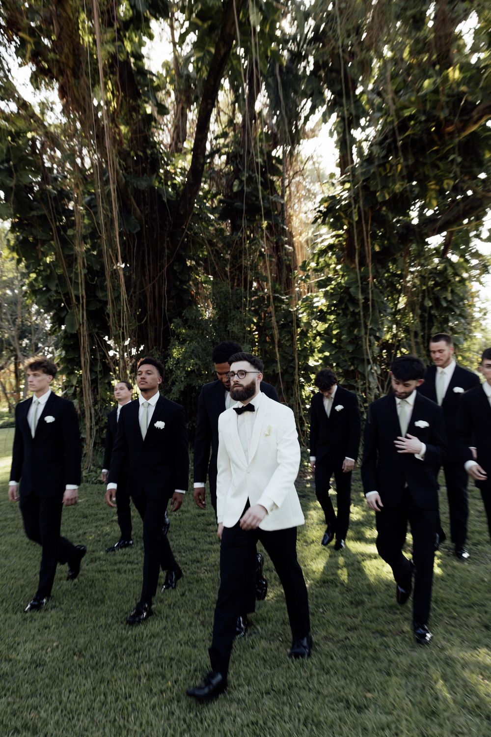 A group of men in formal attire gather under trees for wedding photos.