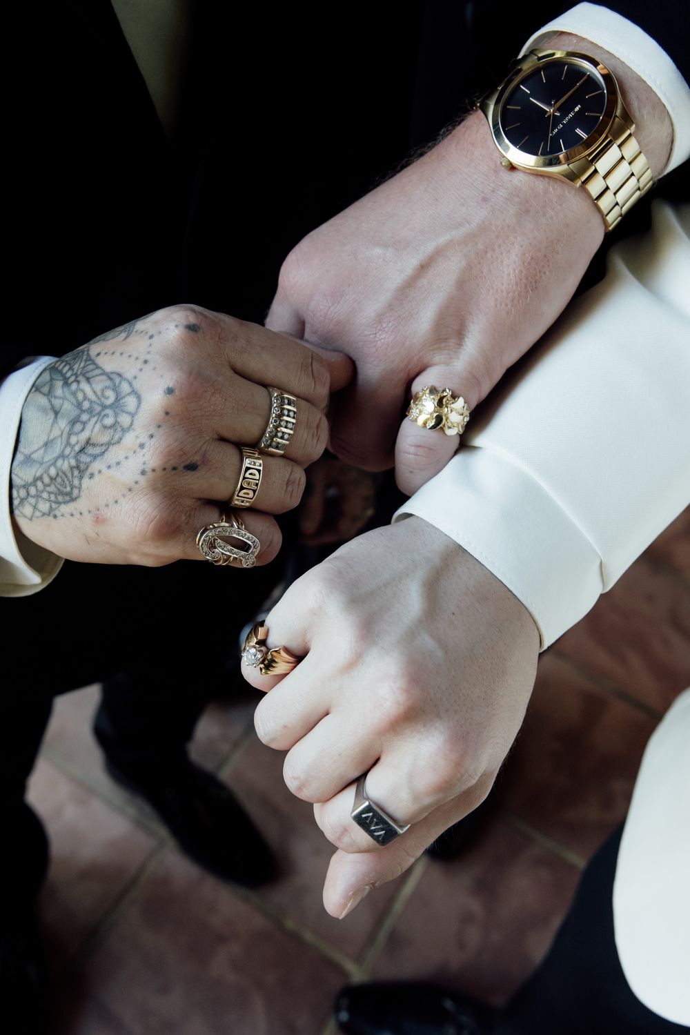 Close-up of hands wearing gold jewelry and watches with visible tattoos against a dark background.