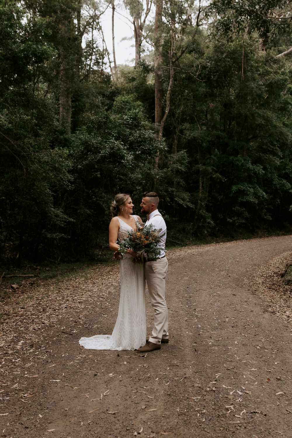 A couple shares an intimate moment on a dirt path surrounded by lush forest greenery during their outdoor wedding.