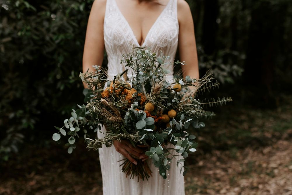 A person in a lace wedding dress holds a rustic bouquet with dried flowers and eucalyptus against a dark background.