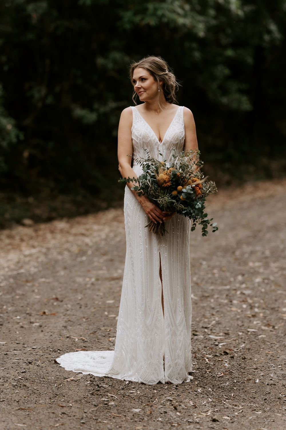 A bride in a flowing lace wedding dress holds a rustic bouquet of wildflowers and eucalyptus on a dirt path.