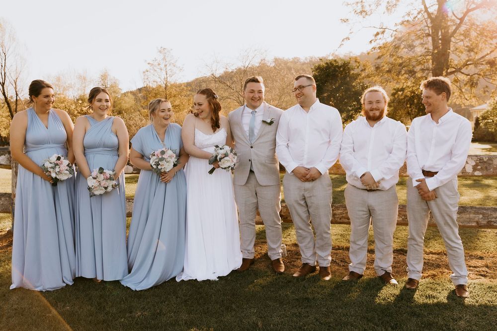 Wedding party in light blue dresses and gray suits poses together outdoors at sunset.