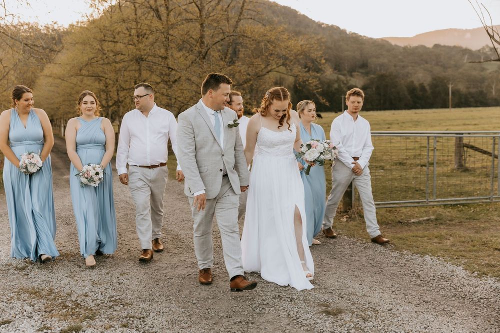 A wedding party in light blue and white attire walks together through a scenic autumn field at sunset.