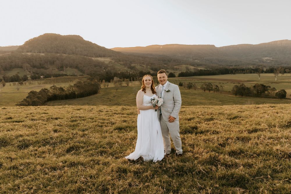 A couple stands together in a golden field during sunset with rolling hills in the background.