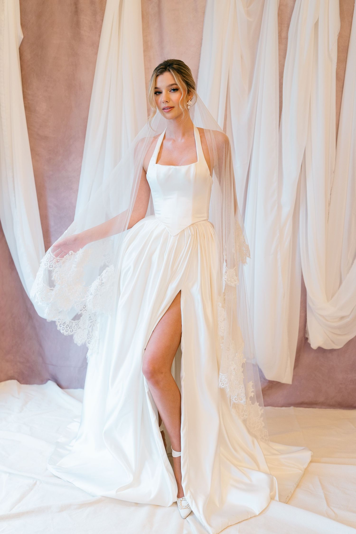 Elegant white wedding dress with halter neckline, thigh-high slit, and flowing veil against draped fabric backdrop.