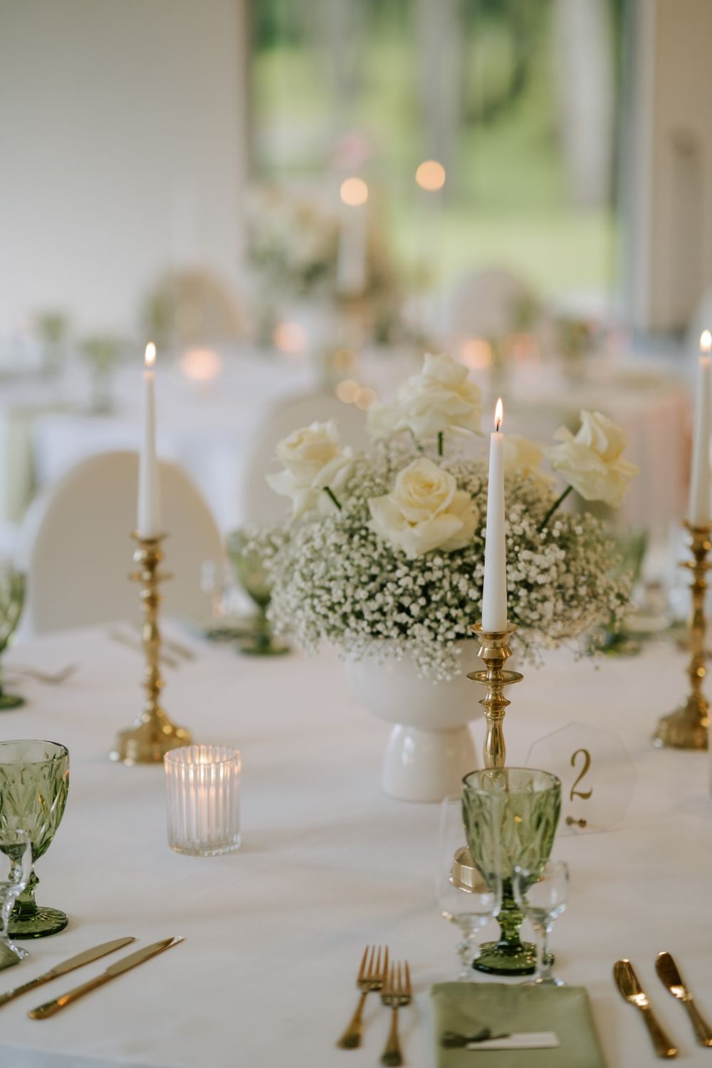 Elegant wedding table setting with white roses, baby's breath, and green glassware with lit candles.