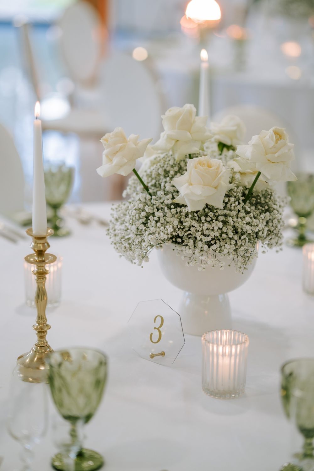 Elegant wedding table settings with white roses, baby's breath centerpieces, and glowing candles on crisp white linens.