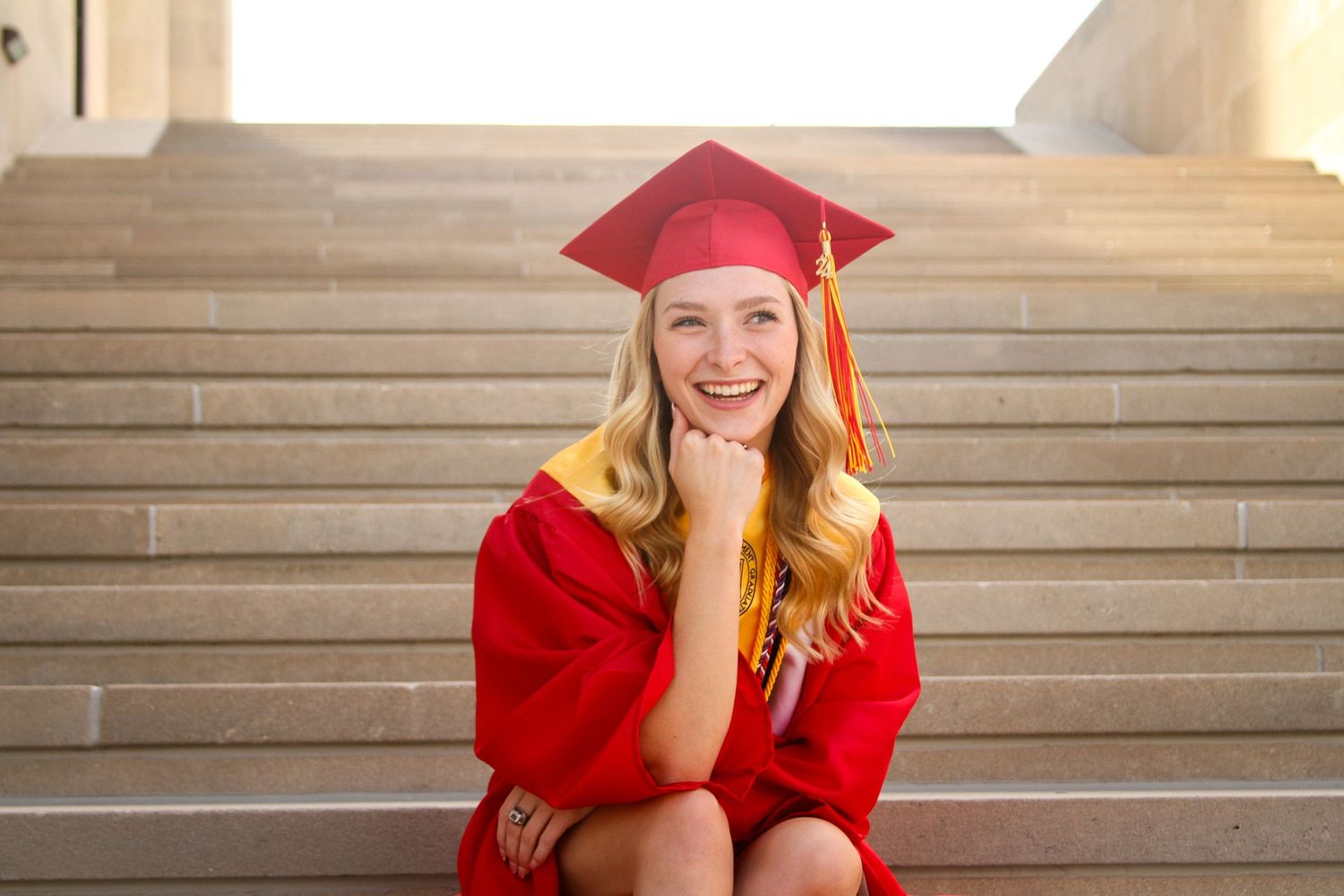 A blonde young woman in a red cap and gown sits on a wide set of stairs, rests her chin on her fist and smiles for her KC senior photos.
