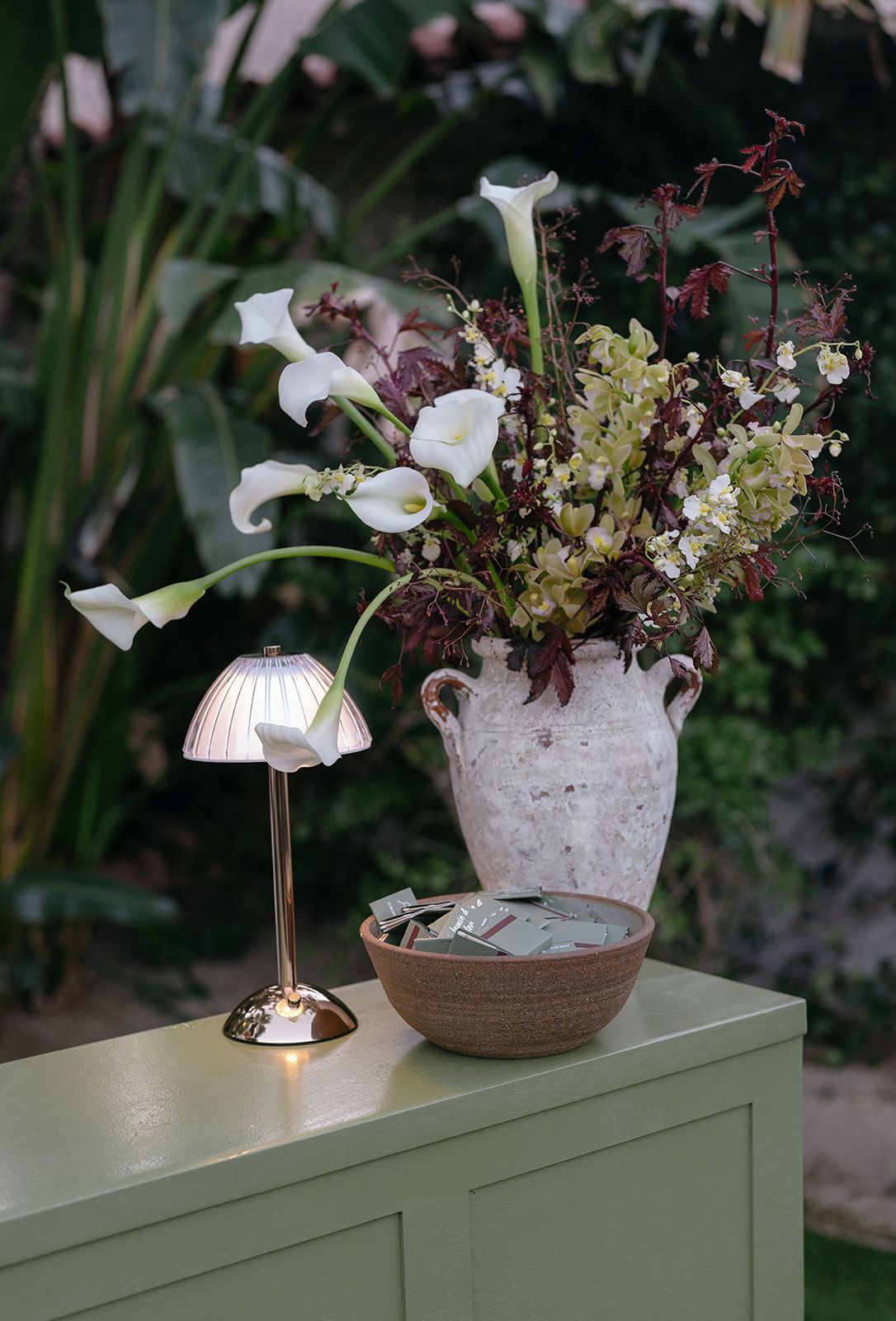 White ceramic planter with delicate flowers sits atop a green surface in a garden setting with tiled roof in background.
