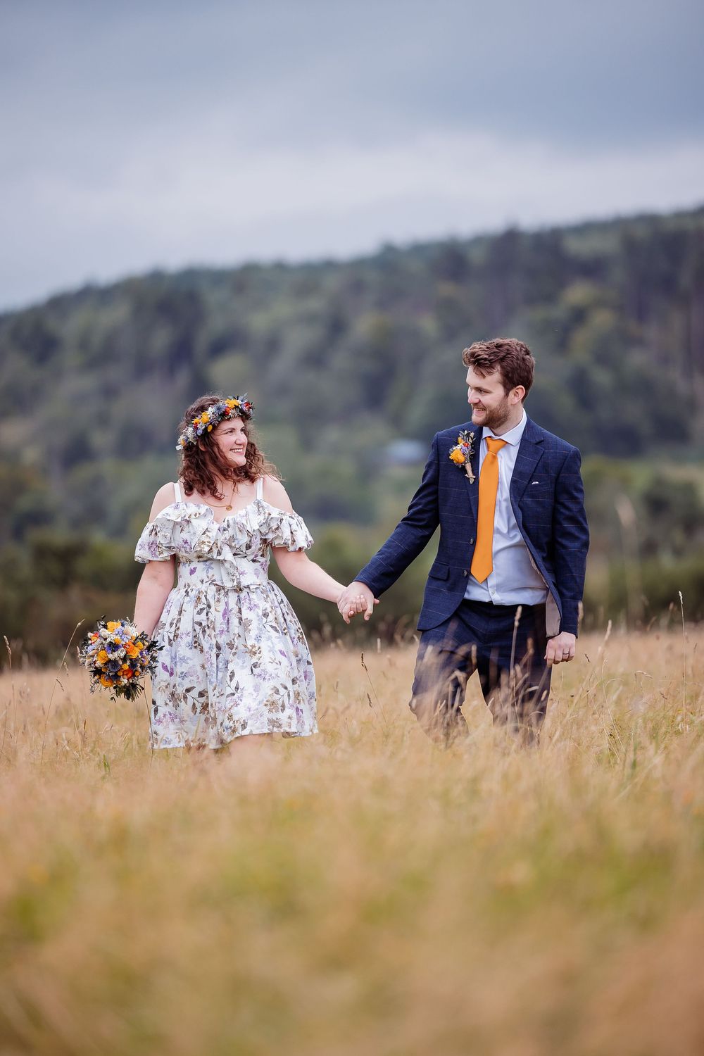 A couple walks hand in hand through a meadow, with the bride wearing a floral dress and groom in navy suit with orange tie.