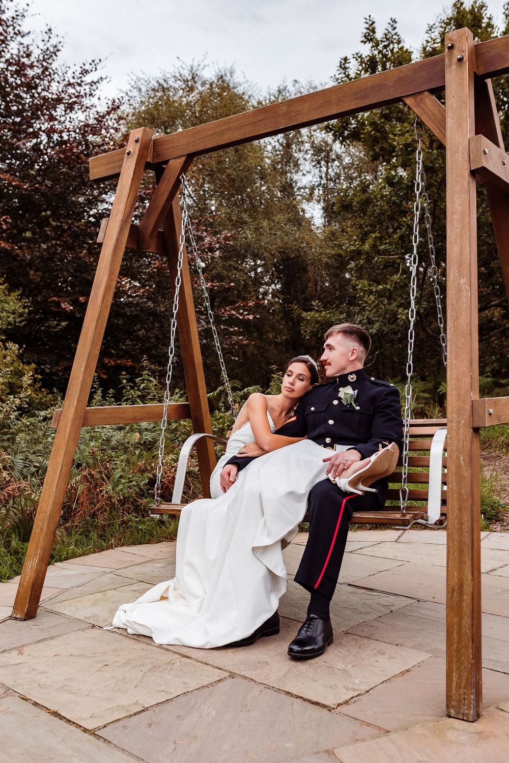 A couple in wedding attire playfully swings together on a wooden swing set in an outdoor setting.